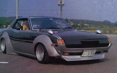 Vintage modified car with wide fenders and sleek design parked on a road with landscape in background.