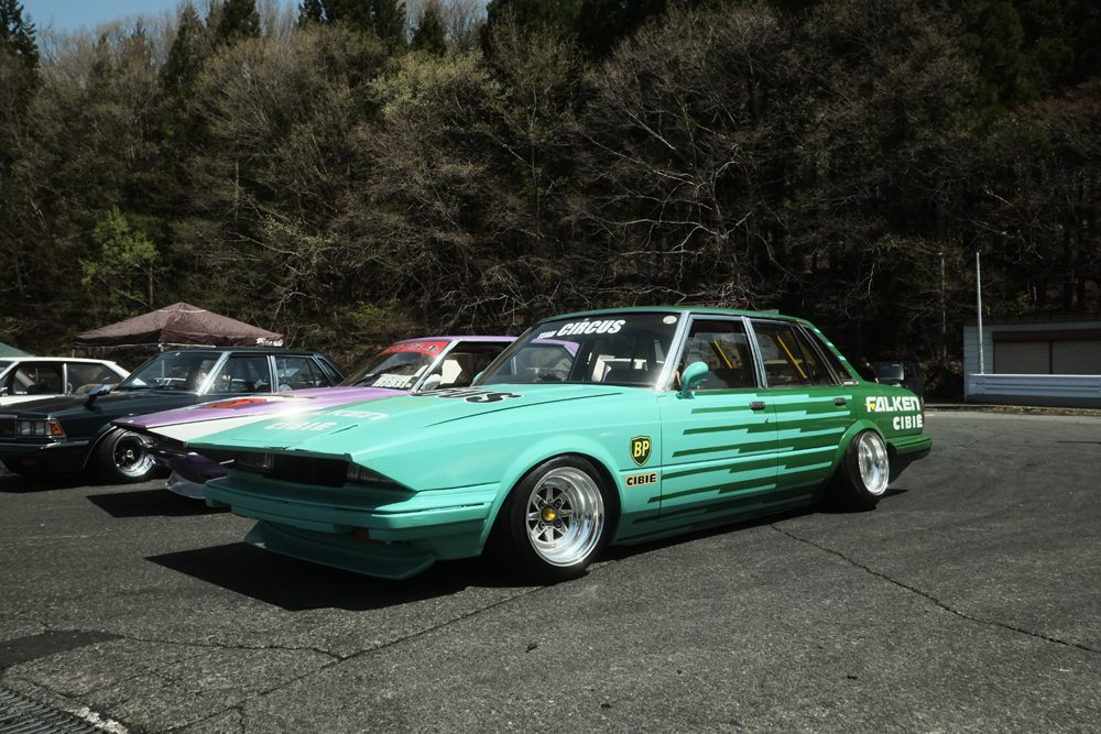 Vintage Toyota GX61 car at a nonsuri car show, featuring racing decals and parked against a wooded backdrop.