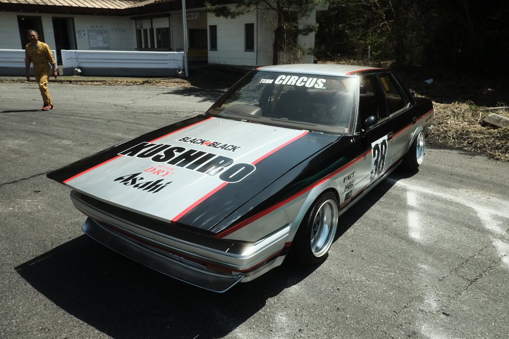 Vintage race car in a parking lot with bold graphics on the hood replicating the japanese gum Black Back, with number 38 on the side.