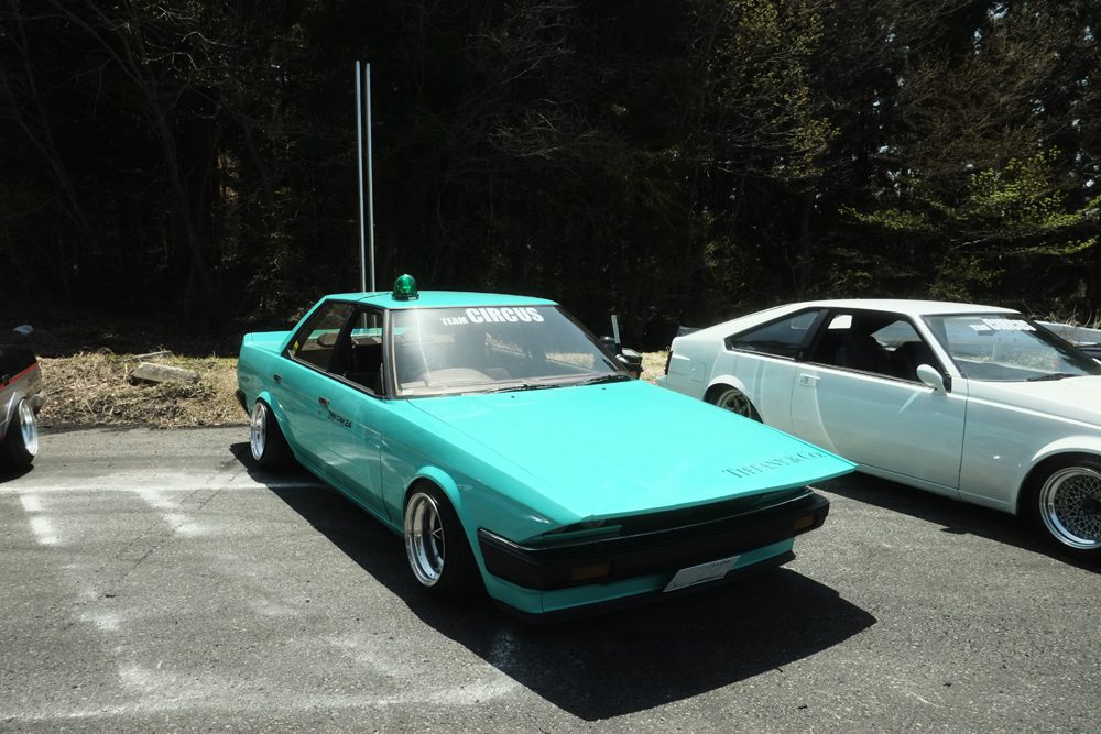 Turquoise vintage car parked outdoors with custom modifications and decals, sunlight casting shadows.
