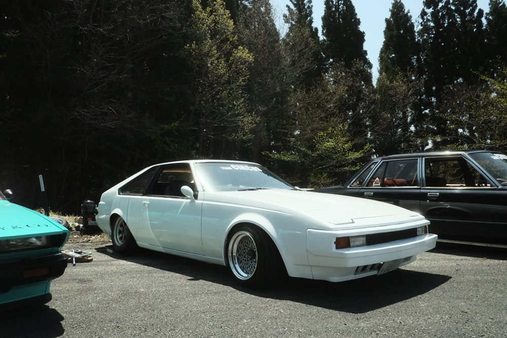 White vintage Toyota Supra or Double XX parked with others under trees on a sunny day.