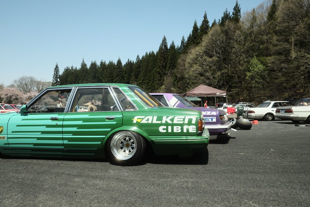 Vintage green race car with Falken logo in a parking lot surrounded by trees and mountains.