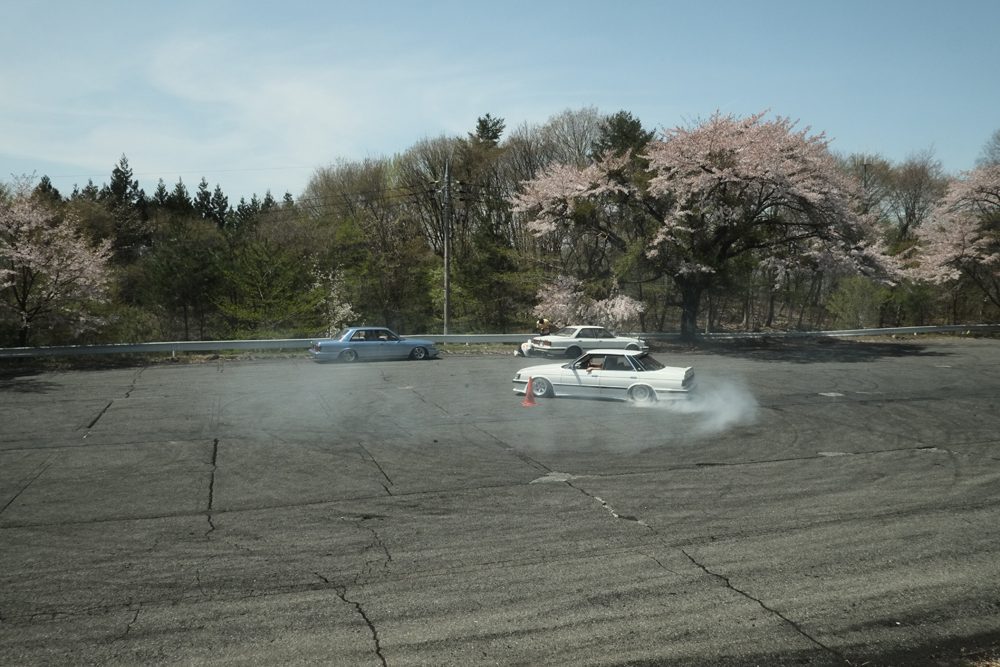Car drifting on a track surrounded by cherry blossoms, producing smoke during a sunny day in a scenic setting.