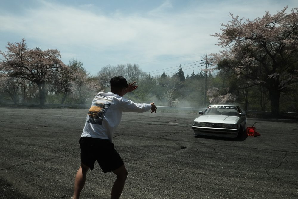 Person directing a drifting car on an empty road with cherry blossom trees in the background.