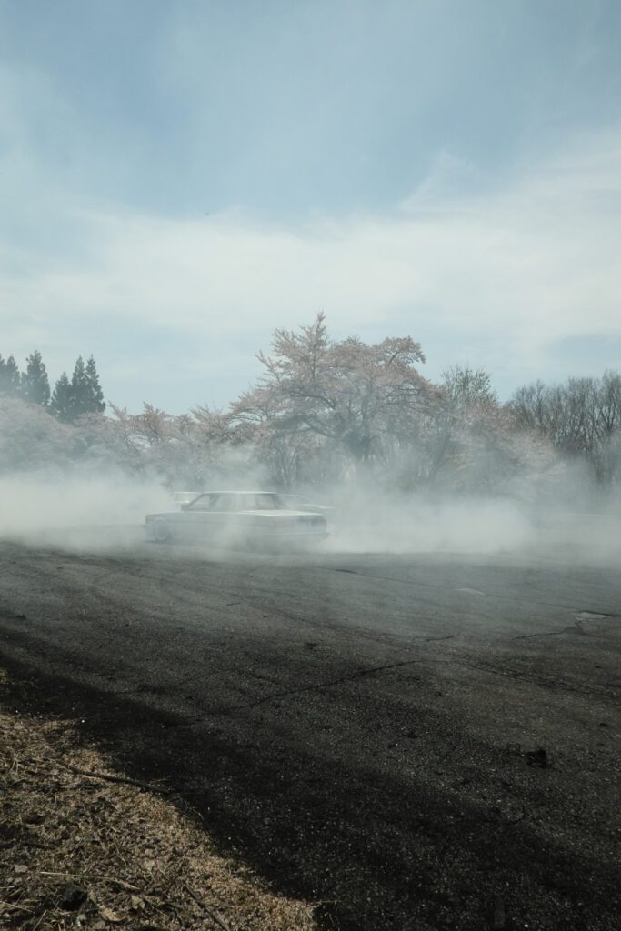 Car drifting through smoky mist on asphalt road with cherry blossom trees in the background.