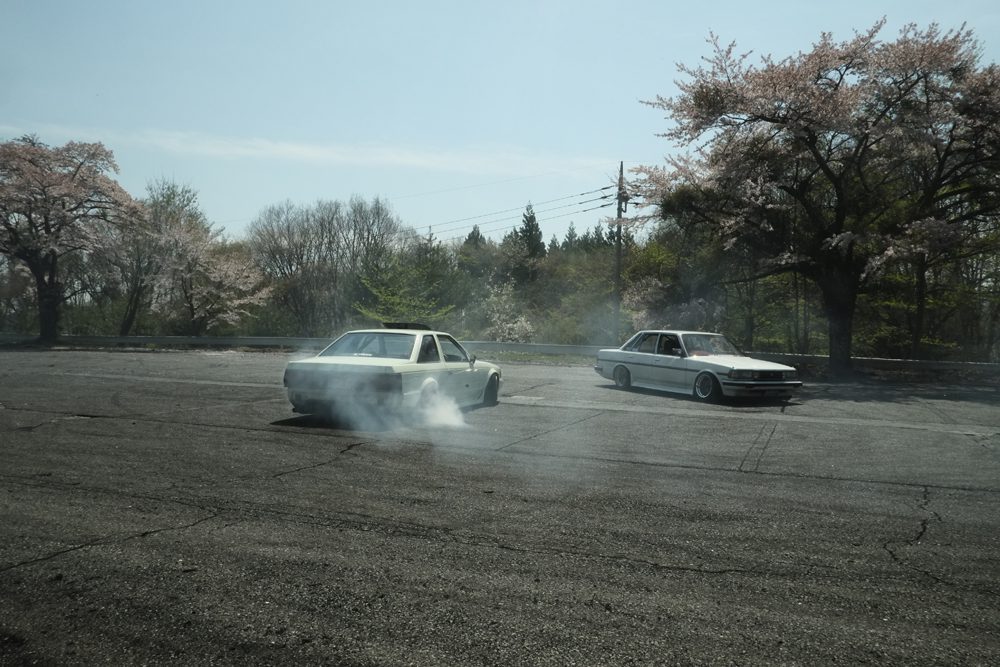 Cars drifting on an open asphalt area with cherry blossom trees in the background, creating smoke clouds.