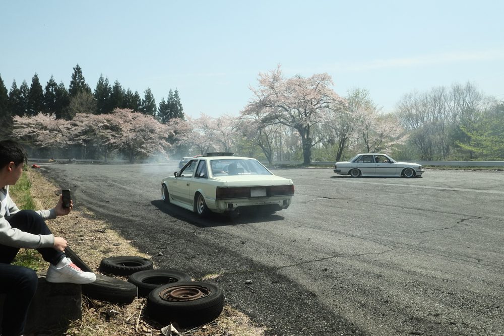 Cars drifting on track near cherry blossom trees, person records with phone.