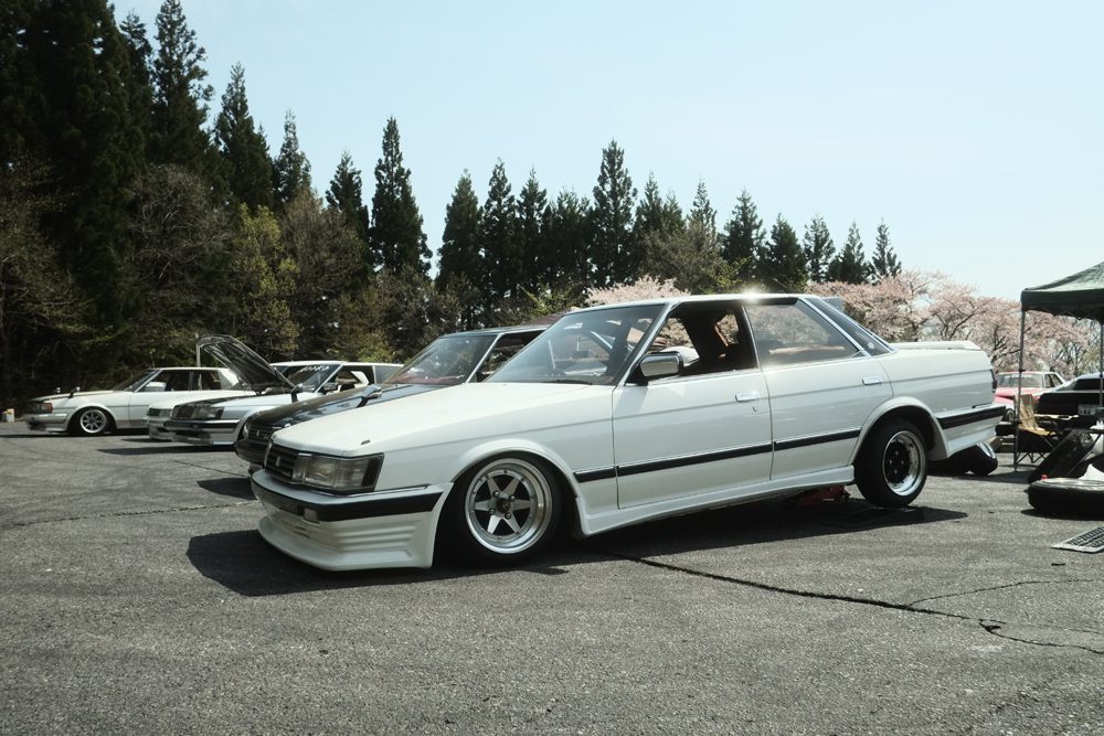White vintage car at an outdoor car show with trees and cherry blossoms in the background.