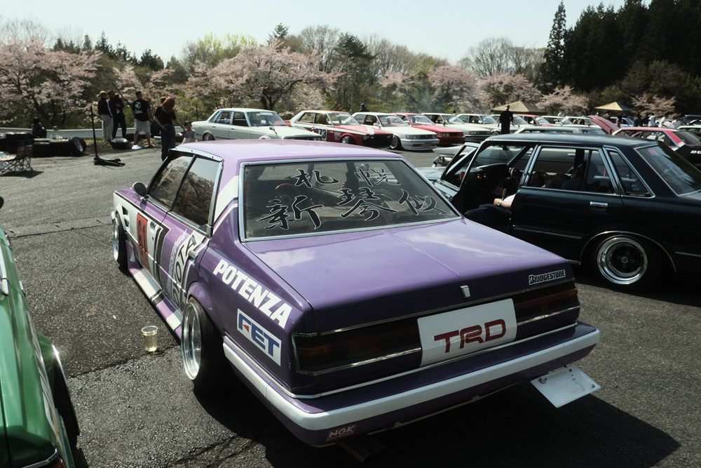 Purple classic car with TRD branding at a car meetup under cherry blossoms.
