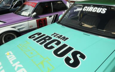 Vintage cars with Team Circus logos, featuring teal and purple designs, parked side by side.
