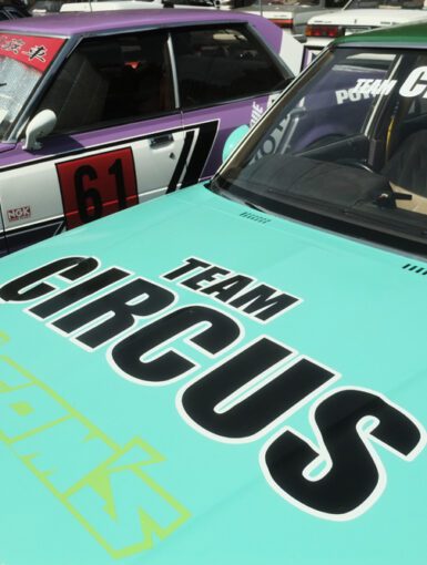 Vintage cars with Team Circus logos, featuring teal and purple designs, parked side by side.