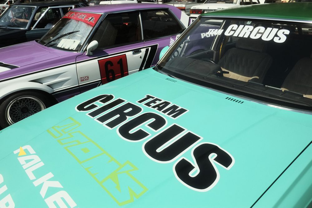 Vintage cars with Team Circus logos, featuring teal and purple designs, parked side by side.