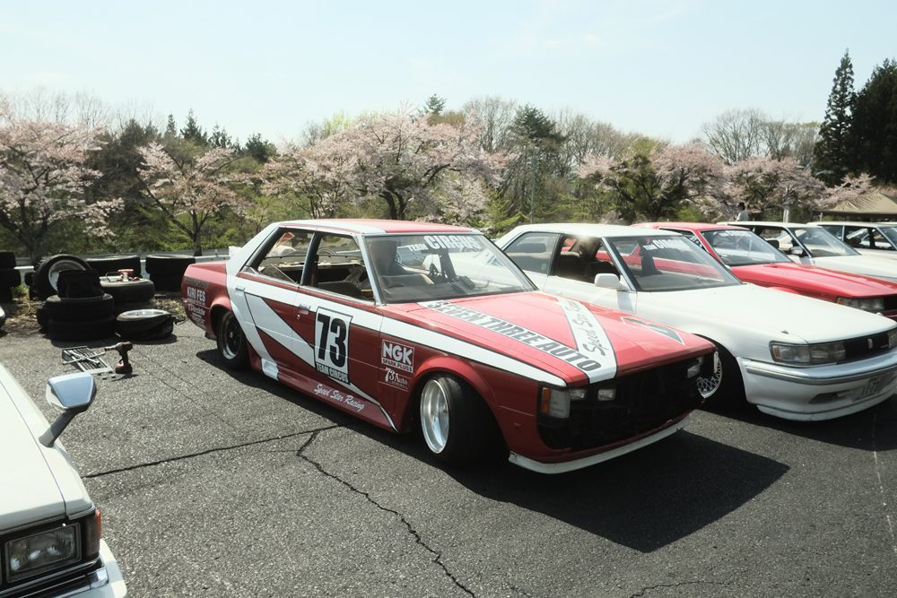 Vintage red race car with 73 on the side, parked near cherry blossom trees in a scenic setting.