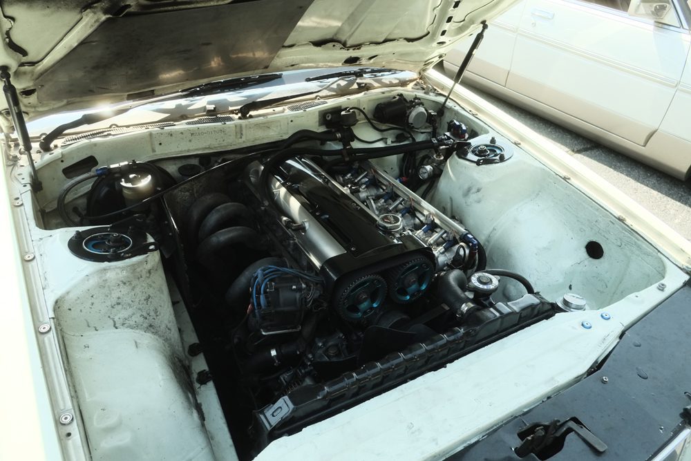 Car engine bay with exposed mechanical components and wiring under an open hood on a sunny day.
