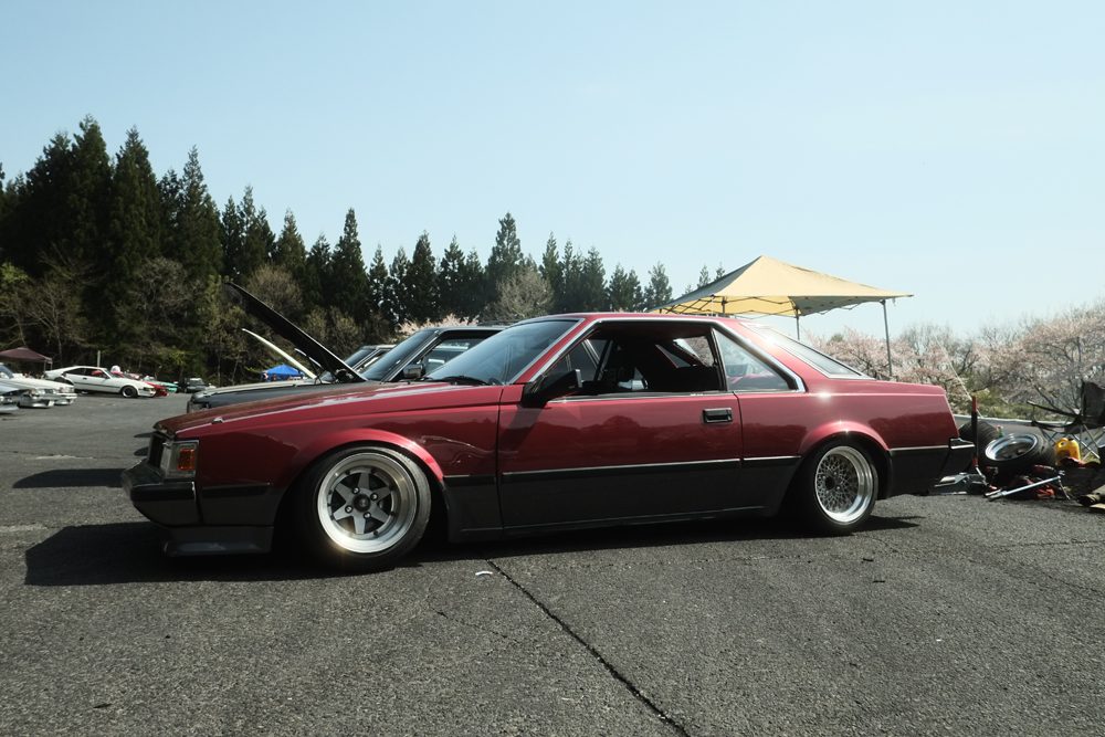 Classic red sports car at an outdoor car meetup, parked under a clear sky with trees in the background.