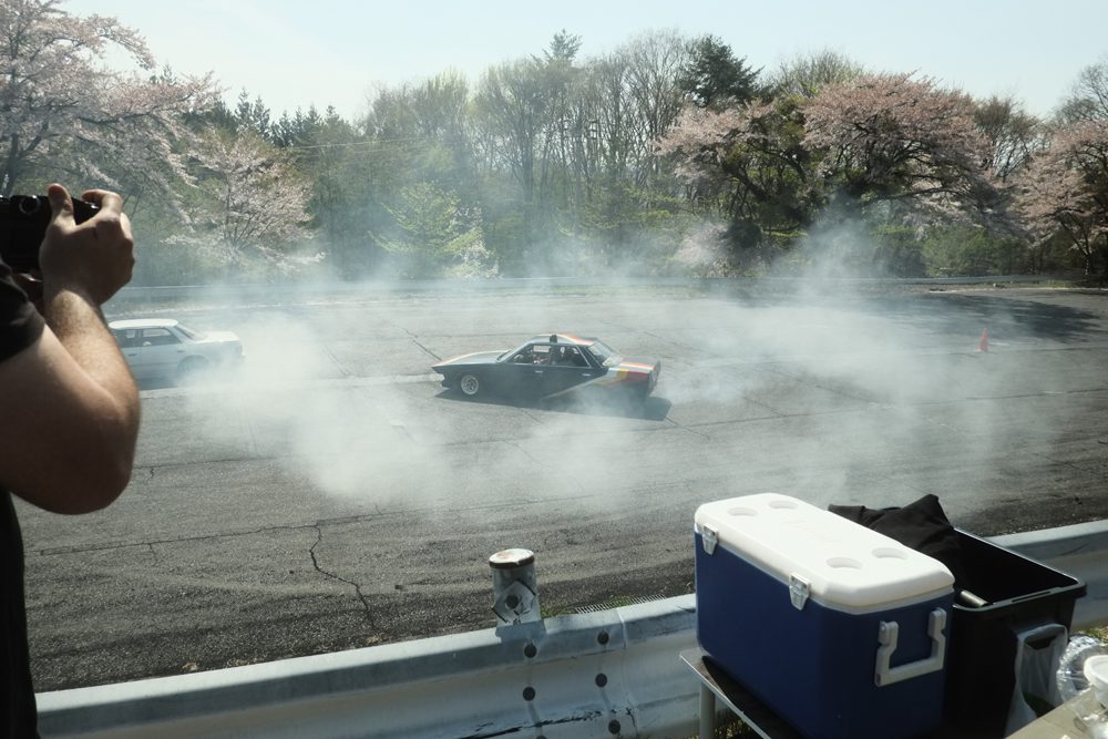 Car drifting on track with smoke, photographed on a sunny day near cherry blossom trees.