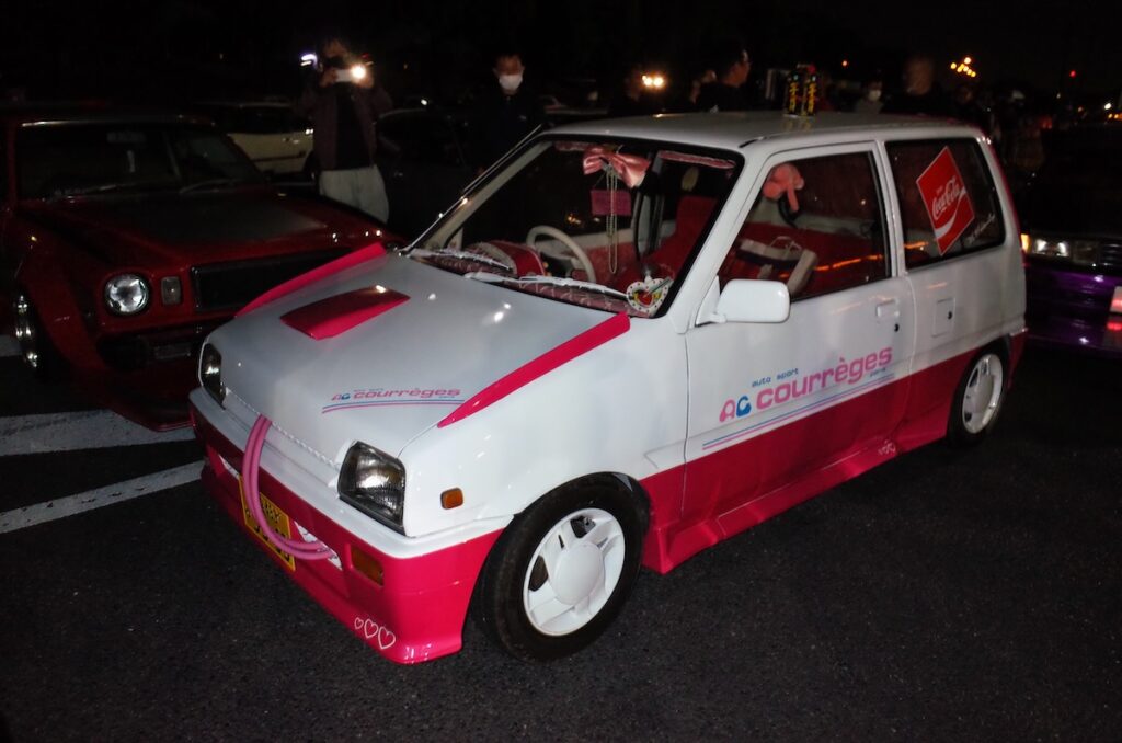 Vintage Daihatsu Mira that is white and pink AC Courrèges car showcased at a night car event with retro decorations.