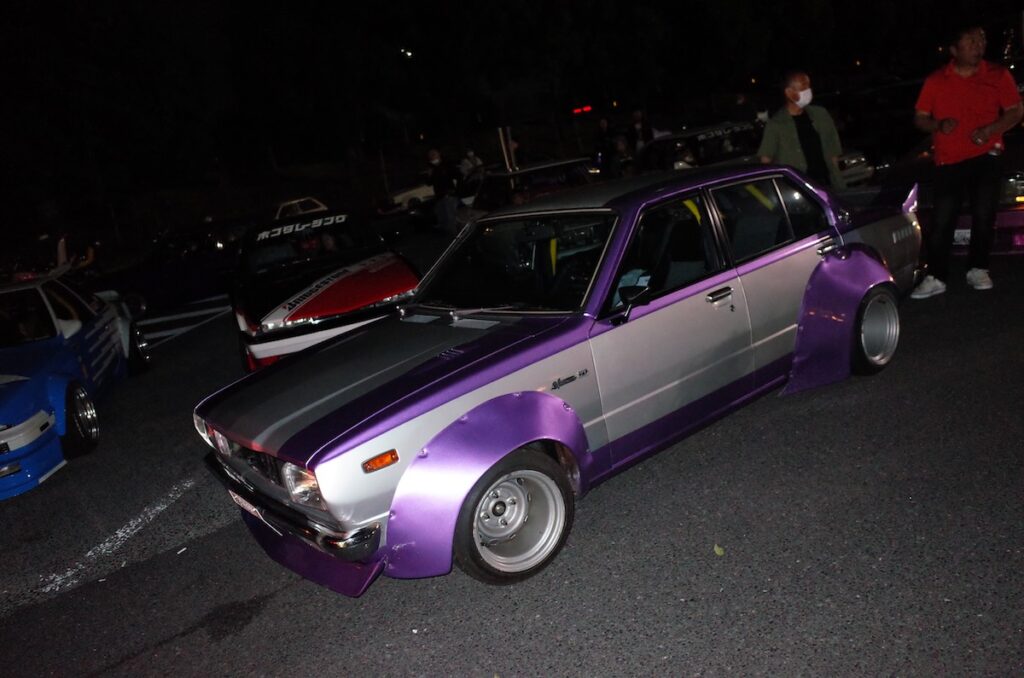 Custom vintage Nissan with purple accents parked at a nighttime car meet, surrounded by enthusiasts.