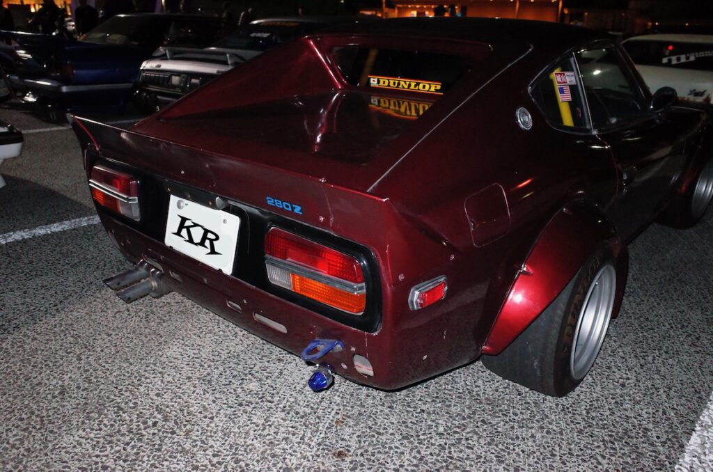 Vintage Datsun 280Z with wide body kit and custom exhaust, showcased at a nighttime car meet.