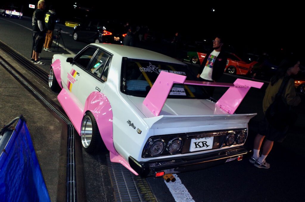 Modified vintage Nissan Skyline with pink accents and spoiler at a night car meet, surrounded by people and other vehicles.