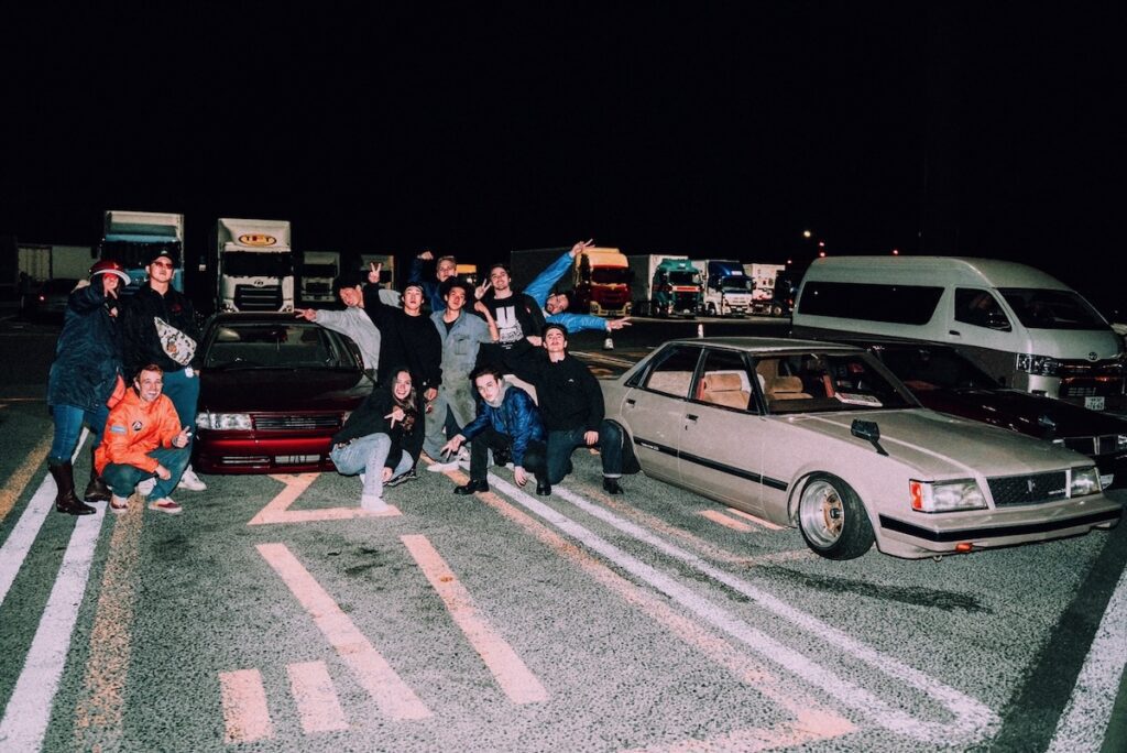 Group gathered at night with vintage cars in a parking lot, celebrating friendship and car culture.