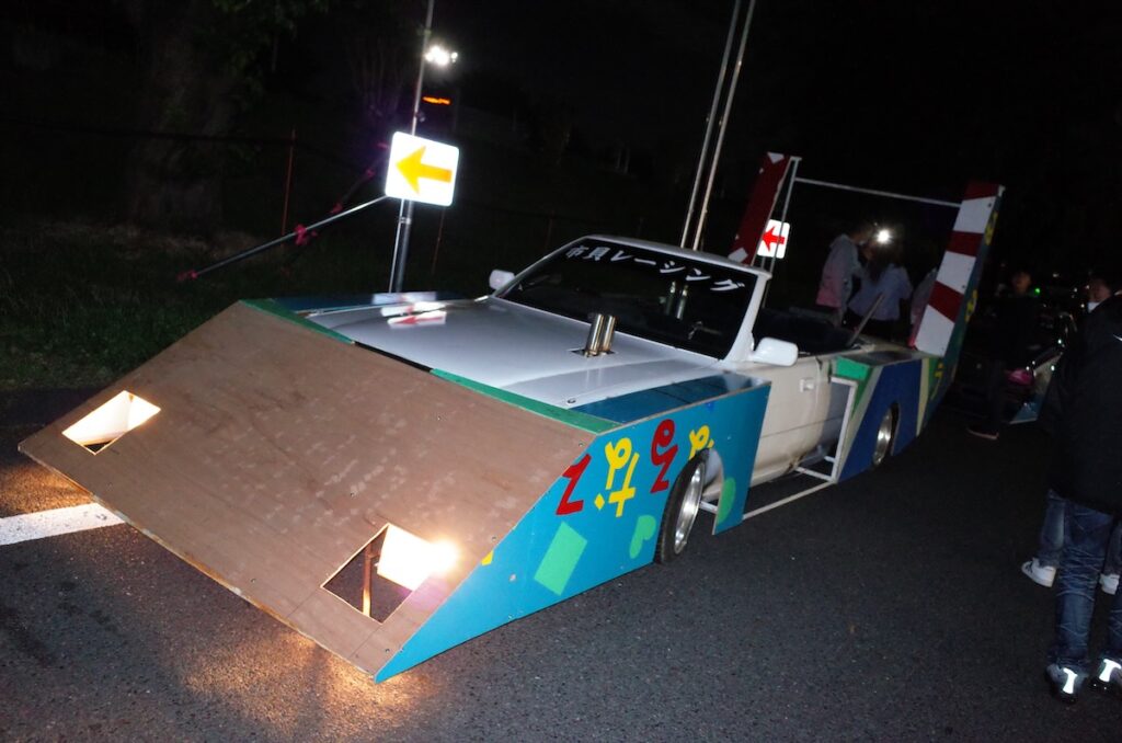 Uniquely decorated car with colorful cardboard design at night, parked under streetlights.