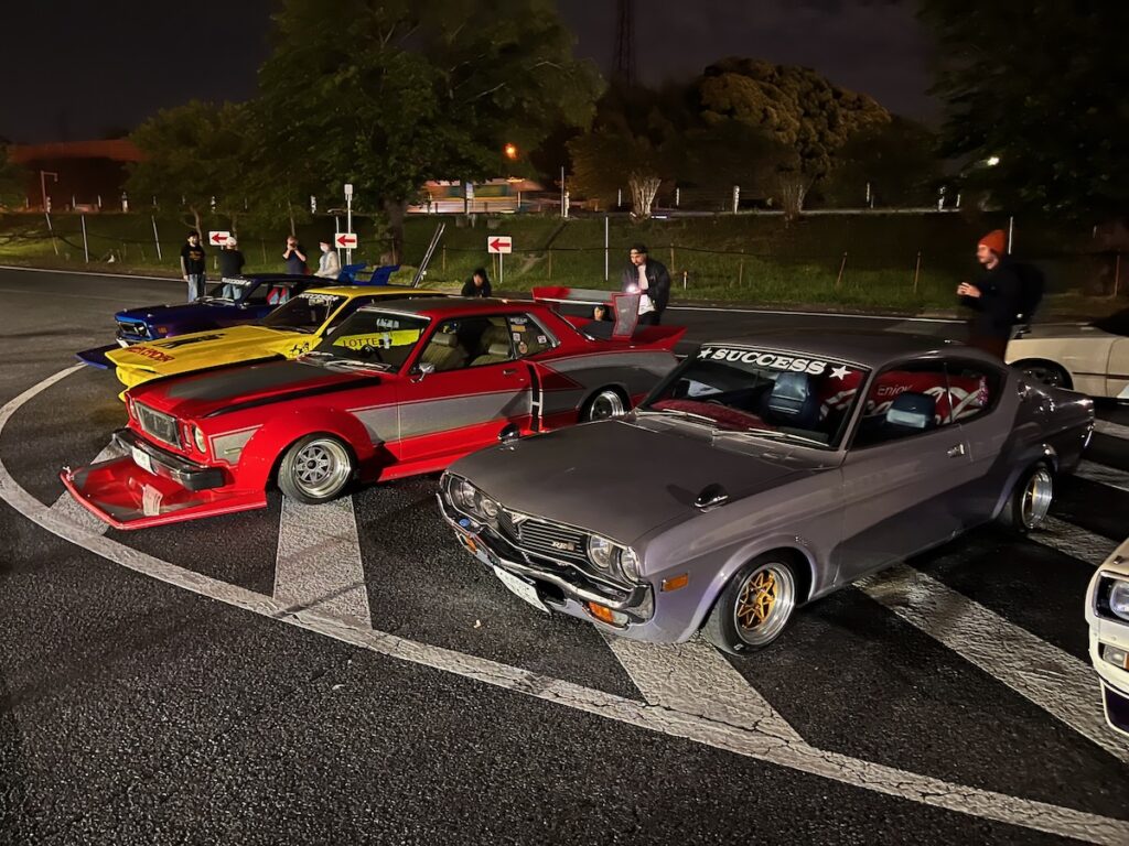 Classic cars lined up in a nighttime parking lot gathering, showcasing vibrant paint and retro styling.