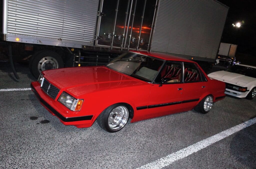 Red vintage car parked at night next to a truck, showcasing sleek design and custom wheels.