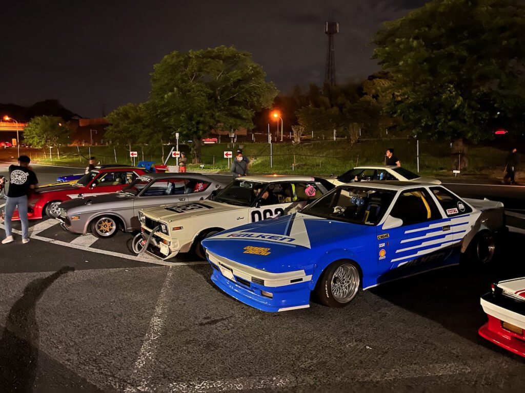 Classic Japanese sports cars parked at night in a lot, featuring blue and white designs, and surrounded by enthusiasts.