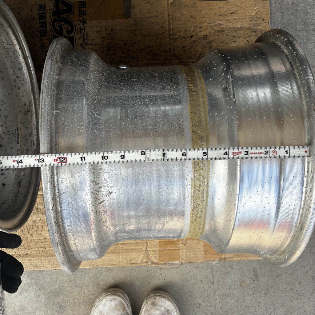 Measuring aluminum wheel rim width with tape measure, clearly showing 16-inch size on a cardboard surface.