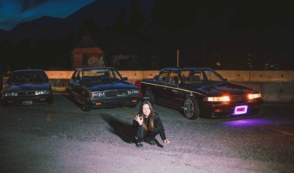 Sandra posing with classic cars at night, illuminated by headlights and purple neon light, in a parking lot.