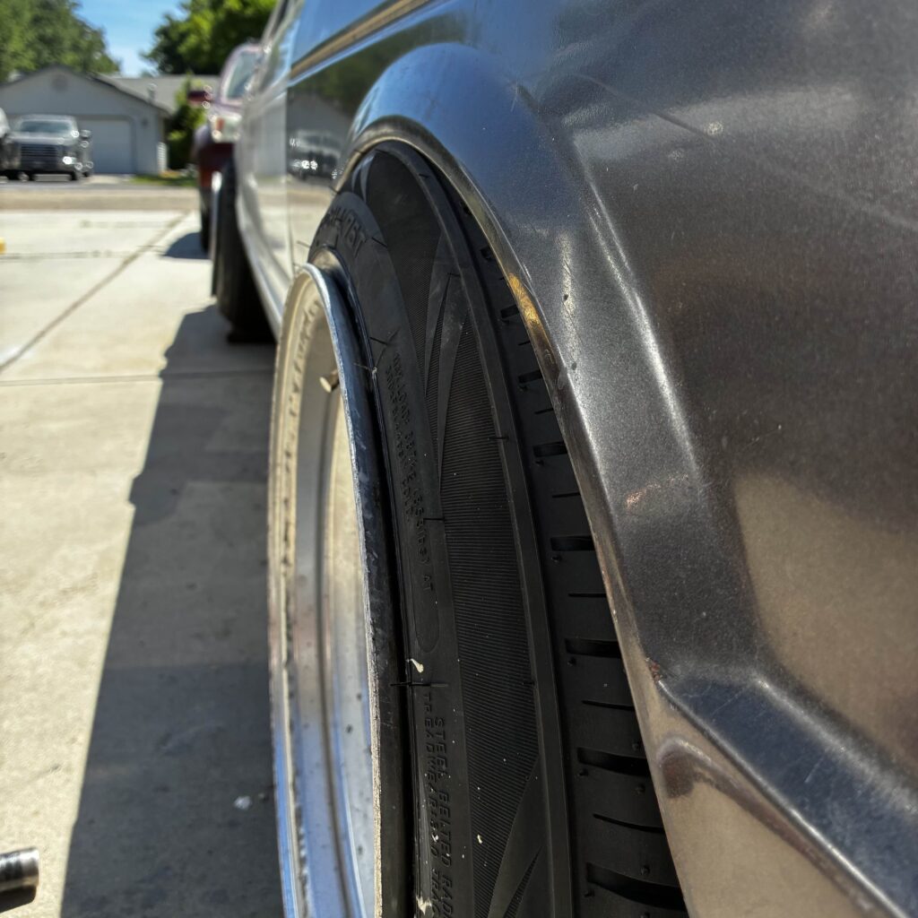 Close-up of a car's rear wheel and fender in a driveway, showcasing a low-profile tire in bright sunlight.