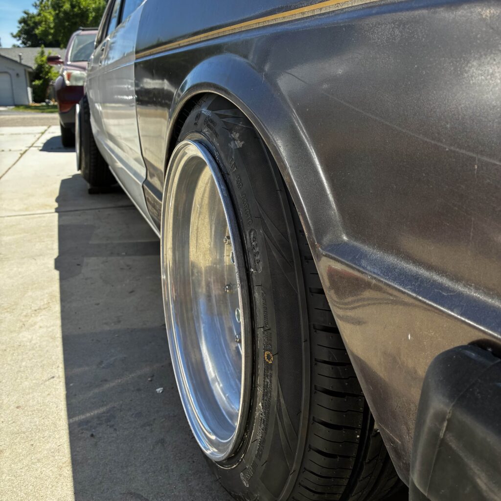 Close-up of a sleek, customized car wheel with wide rims and tires in a driveway on a sunny day.