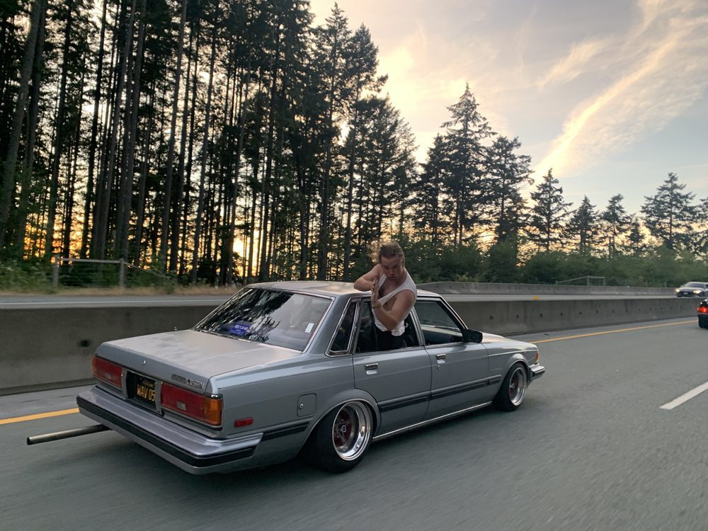 Person leans out of classic silver car on a scenic forested highway during sunset, capturing adventurous road trip.