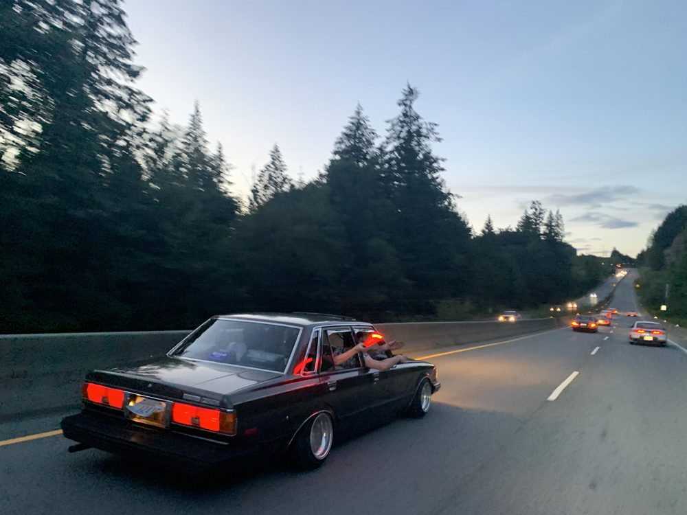 Vintage car on a scenic highway at dusk, surrounded by trees and other vehicles, with a passenger enjoying the ride.