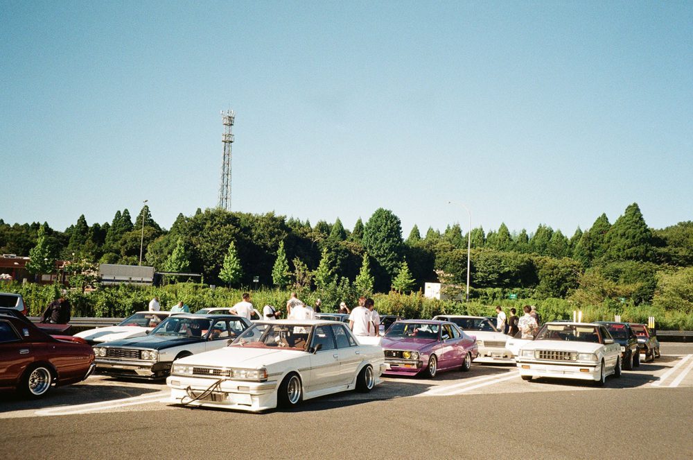 Classic cars gathering at a scenic parking lot with people and trees in the background under a clear blue sky.