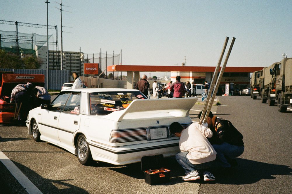 Two people modify a white car with a large spoiler at a busy gas station, surrounded by trucks and other vehicles.