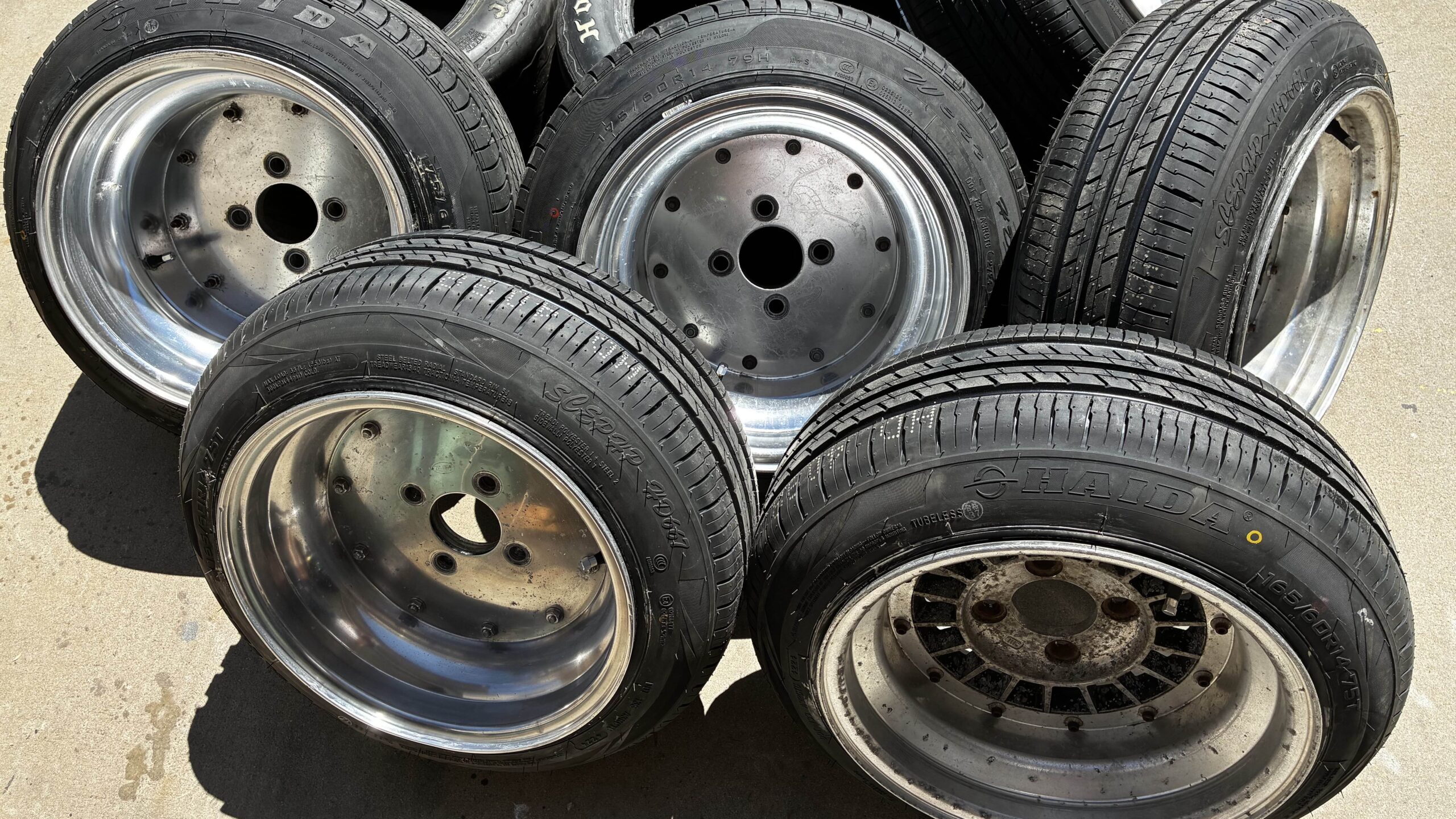 A collection of car rims and tires stacked on concrete in sunlight, showcasing different tread patterns and styles.