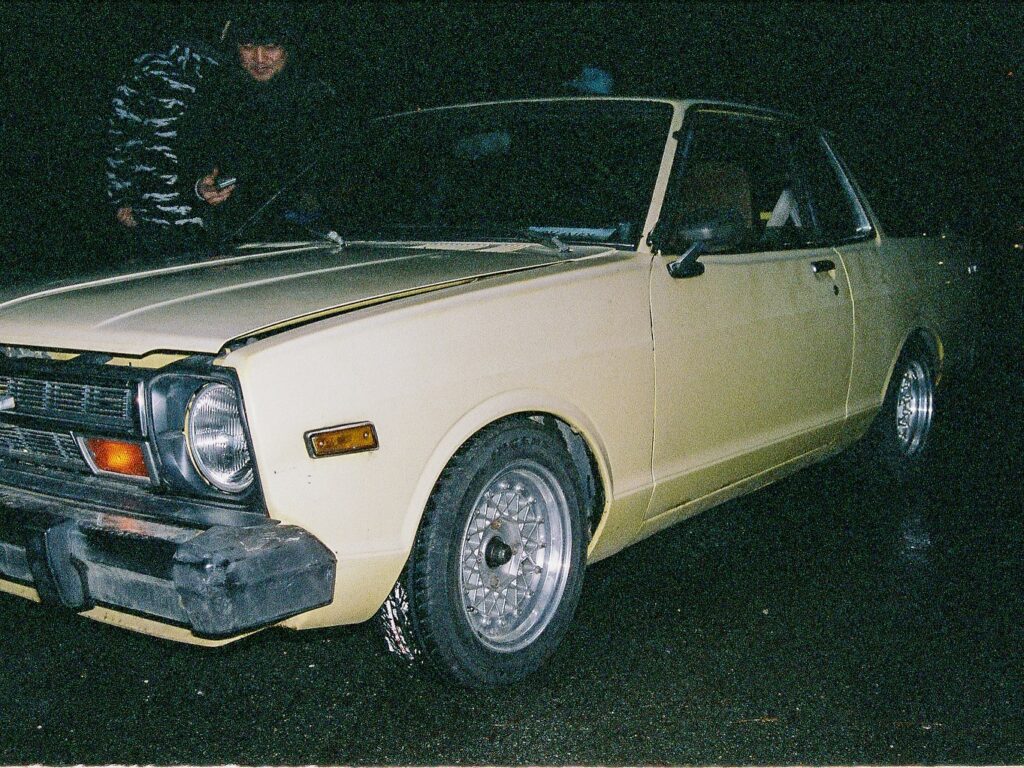 Vintage yellow car at night, with a person in a camo jacket nearby. Classic design with round headlights.