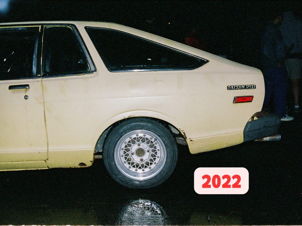 Vintage Datsun 210 in yellow parked at night, visible wear on rear wheel arch, water reflections on pavement, 2022.