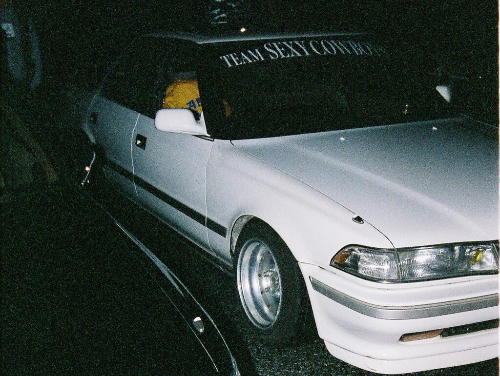 White car with Team Sexy Cowboys windshield banner, parked at night, featuring sporty wheels and a sleek body design.