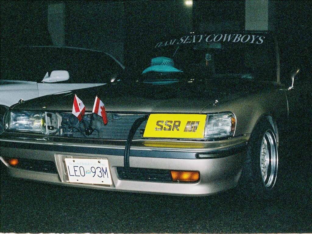 Vintage car with two Canadian flags, SSR plate, and TEAM SEXY COWBOYS windshield decal, parked at night.