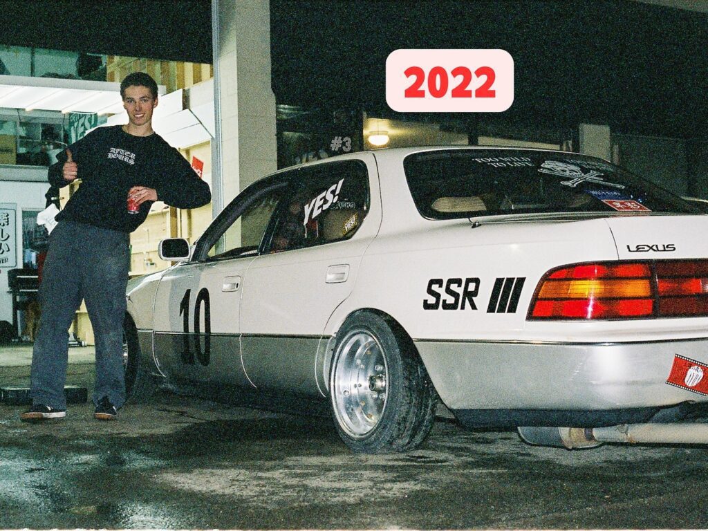 Person posing with a modified Lexus car, displaying YES! and SSR III decals at night, with a 2022 graphic.