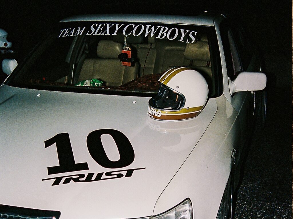 Racing car with Team Sexy Cowboys and TRUST 10 decals, helmet on hood.