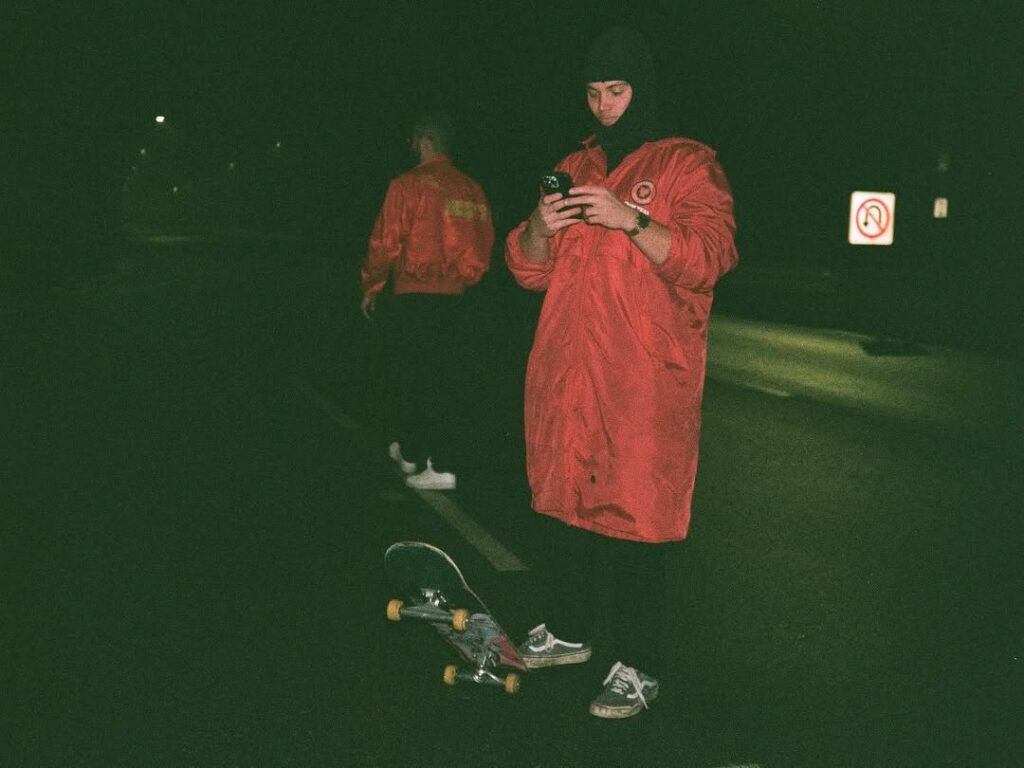 Person in red coat uses phone beside skateboard at night on a dimly lit street.
