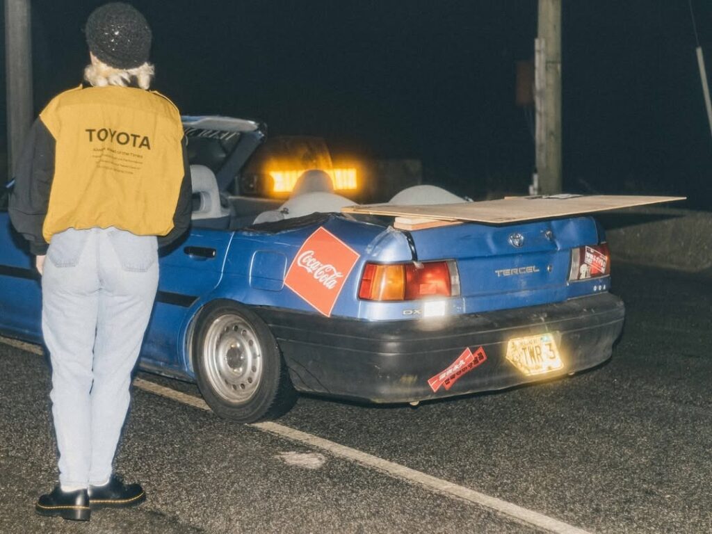 Person standing by a blue Toyota Tercel with Coca-Cola logo, parked at night on a road with wooden board on trunk.