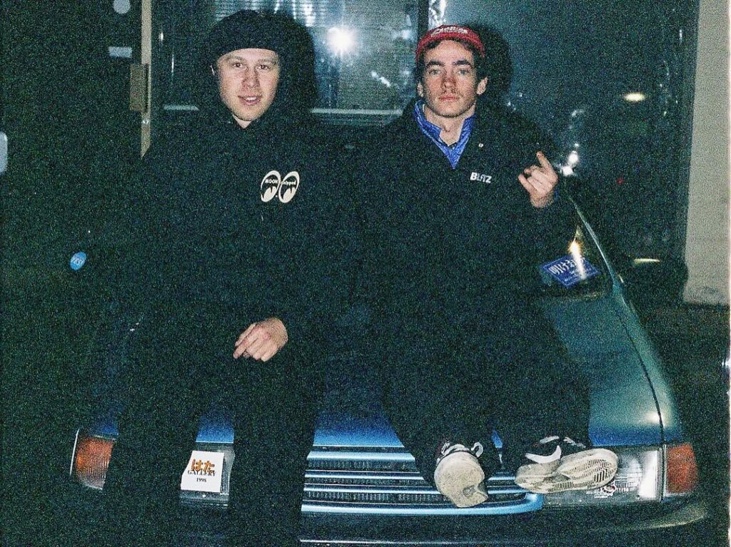 Two people sitting on a car hood at night, wearing casual clothing and hats, with a flash in the background.