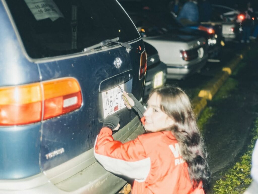Person in a red jacket inspecting a blue car's license plate in a parking lot at night.
