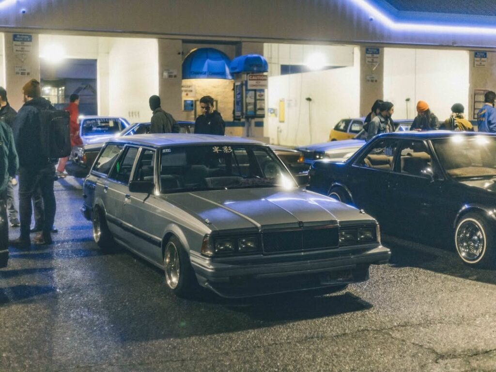 Vintage cars gather at a nighttime car meet, surrounded by enthusiasts under neon lights.