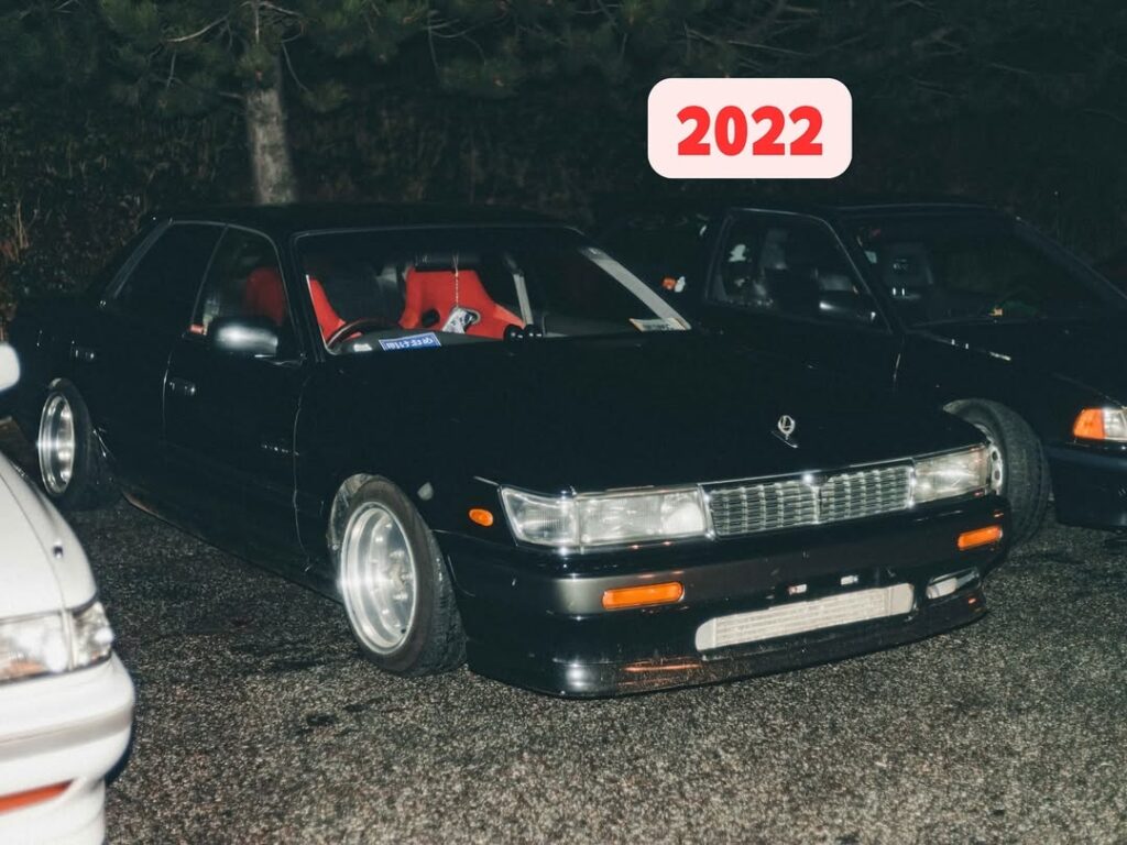 Black vintage car with red interior parked at night, labeled 2022, surrounded by other vehicles.